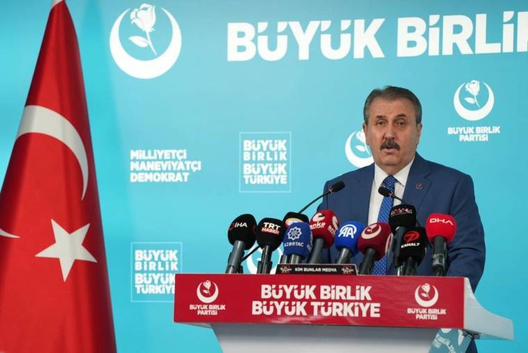 Kürsüde konuşan bir kişi, mikrofonlar önünde duruyor; arkasında parti logoları ve Türk bayrağı bulunuyor.