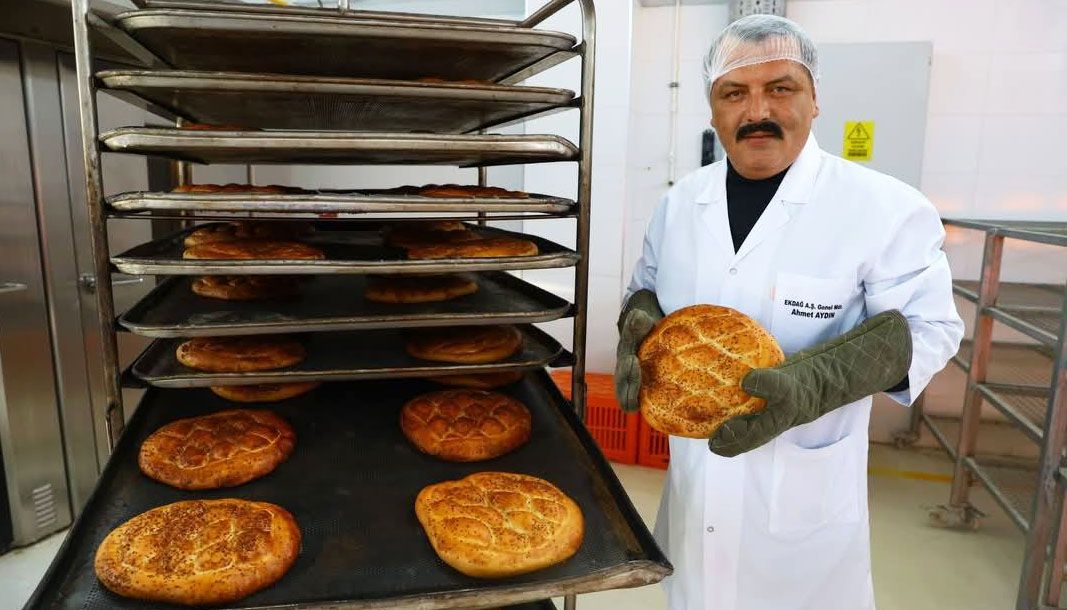 Fırın çalışanı, eldivenlerle bir ramazan pidesi tutarken yanında pidelerle dolu tepsi rafı duruyor.