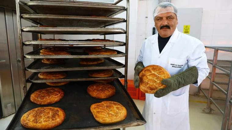 Fırın çalışanı, eldivenlerle bir ramazan pidesi tutarken yanında pidelerle dolu tepsi rafı duruyor.