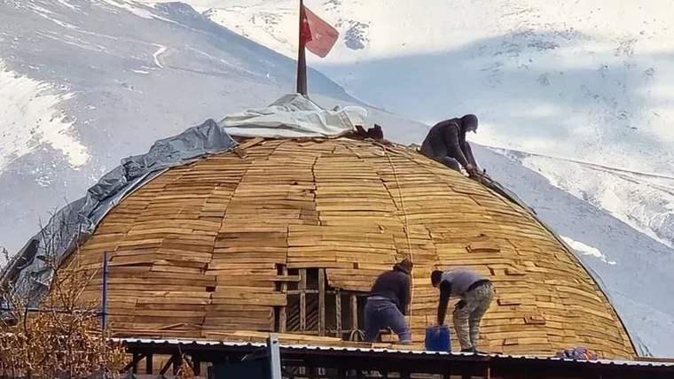 Karla kaplı dağların önünde, Türk bayrağı bulunan kubbeli çatıda dört kişi çalışma yapıyor.