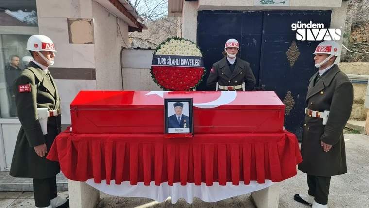 Türk bayrağına sarılı tabut, yanında askerler duruyor ve bir çiçek aranjmanı var.