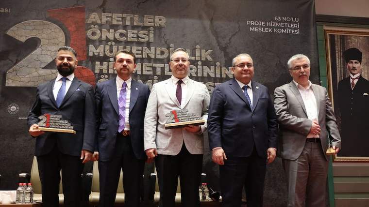 Beş erkek, ellerinde plaketlerle bir salonun sahnesinde yan yana duruyor; arkalarında etkinlik yazısı bulunuyor.