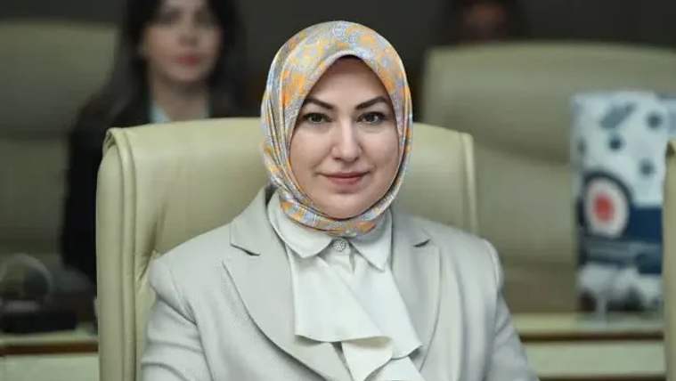 Krem rengi bir ceket giymiş, başında desenli bir başörtüsü olan bir kadın, oturduğu koltukta gülümsüyor.