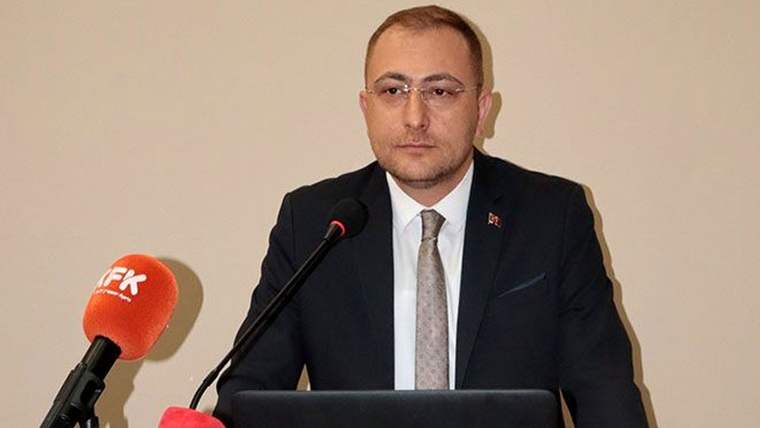 Kürsüde konuşan bir kişi, önünde mikrofon ve bilgisayar ekranı var.
