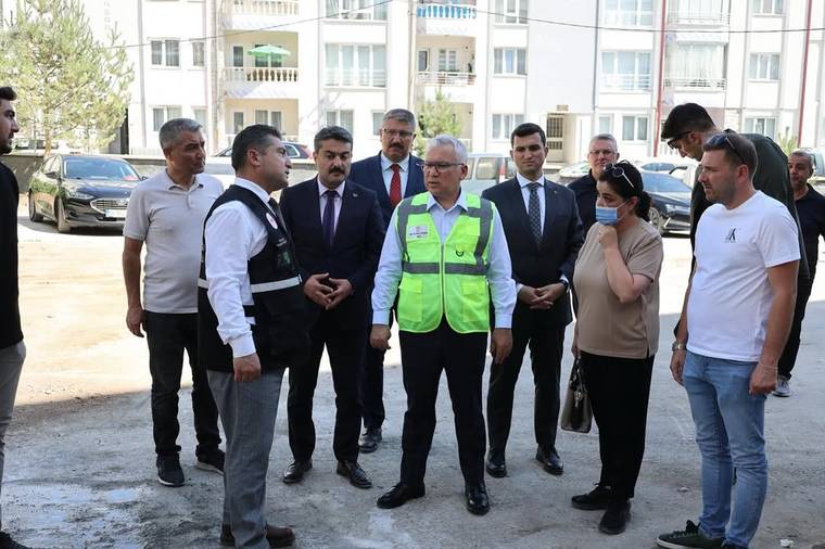 Açık alanda, baretli görevlinin çevresinde duran bir grup kişi ve arkada apartman binaları görülüyor.
