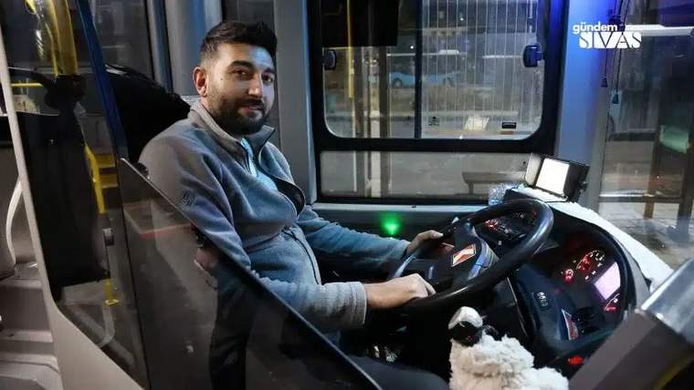 Otobüsün sürücü bölümünde oturan bir kişi, direksiyon ve gösterge panelinin yanında kameraya bakıyor.