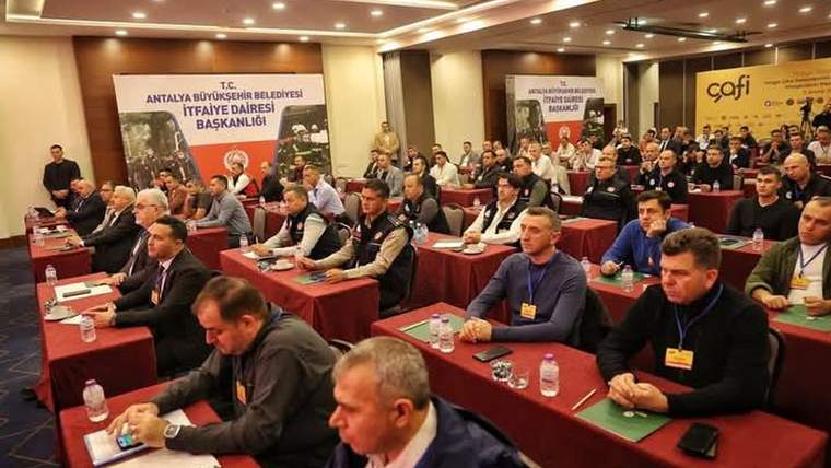 Toplantı salonunda sıralar halinde oturan kalabalık, önlerinde su şişeleri ve not defterleriyle görülüyor.