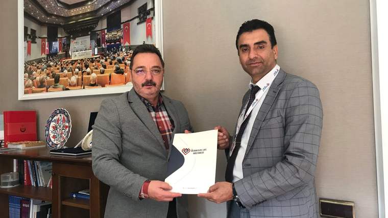 Bir ofiste iki erkek, üzerinde logo bulunan bir belgeyi birlikte tutuyor; arkada toplantı fotoğrafı ve Türk bayrakları var.