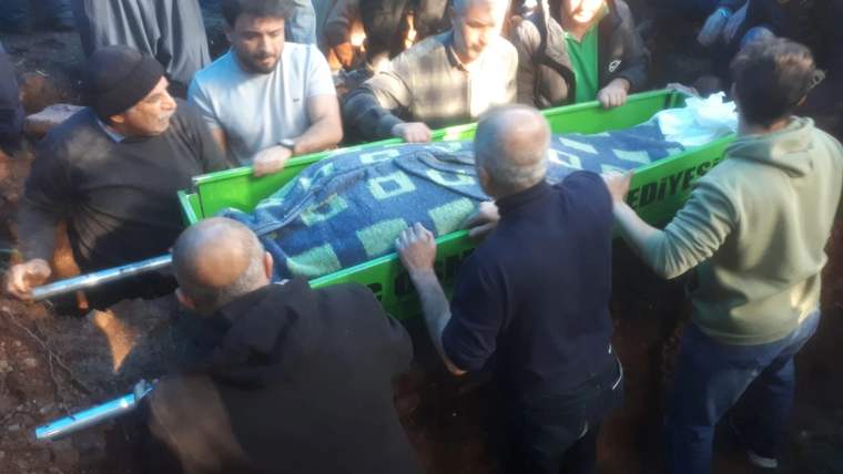 Kalabalık bir grup, üzeri örtülü yeşil bir tabutu ormanlık alanda taşırken görülüyor.