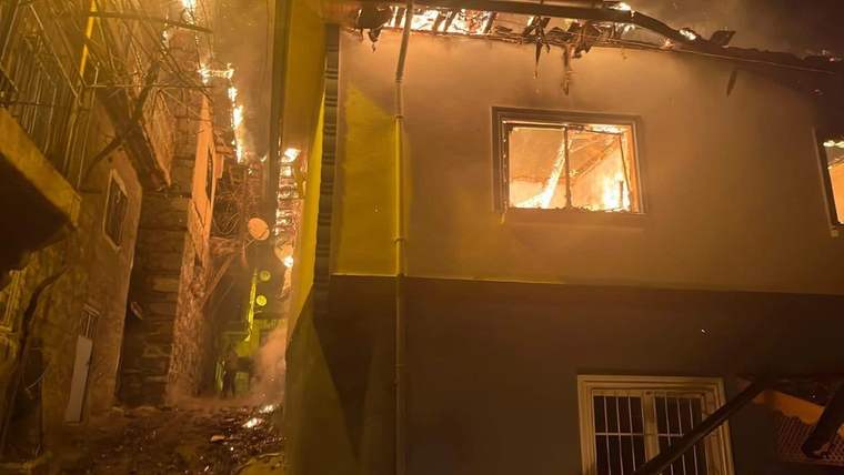 Alevlerin sardığı binanın çatısı ve pencereleri görünürken, dar sokakta yoğun duman yükseliyor.