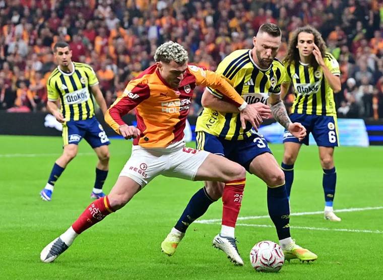 Futbol sahasında sarı-lacivertli ve turuncu-kırmızılı formalı oyuncular top için mücadele ediyor.