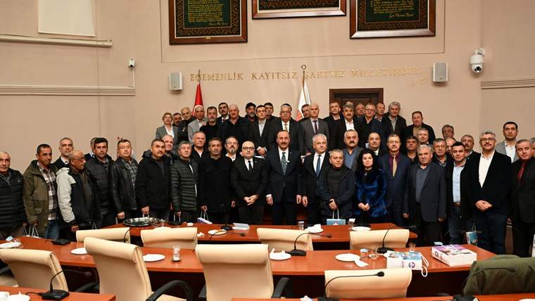 Toplantı salonunda, Türk bayrakları ve duvar yazısı önünde çok sayıda kişi birlikte poz veriyor.