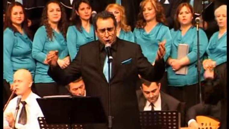 Mikrofon önünde konuşan bir erkek, arkasında turkuaz giyimli kadın korosu ve oturan müzisyenler bulunuyor.