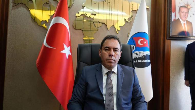 Makam koltuğunda oturan bir kişi, Türk bayrağı ve belediye amblemli bayrakların önünde görülüyor.