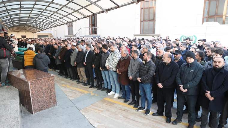 Üzeri yeşil örtüyle kaplı tabutun yanında, kapalı alanda saf tutarak bekleyen kalabalık