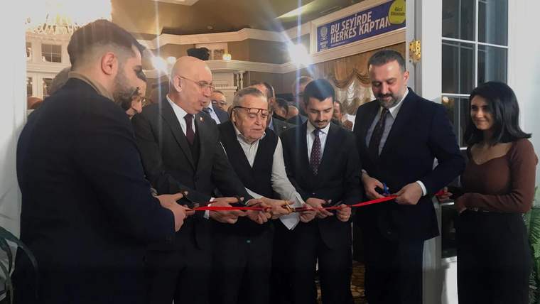 Bir grup insan, bir binanın girişinde kırmızı kurdeleyi makaslarla keserken görülüyor.