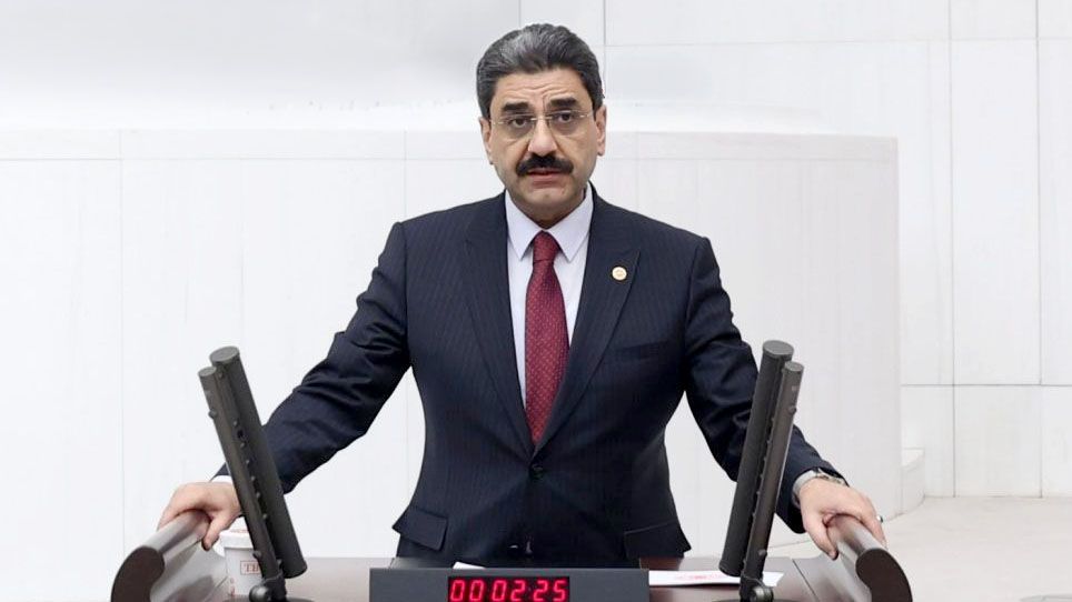 Kürsüde konuşan bir kişi, arkasında beyaz bir duvar var.