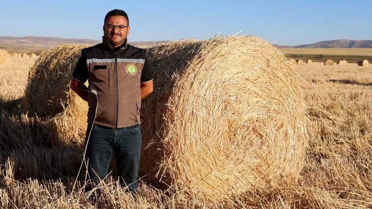 Bir adam, arkasında büyük rulo balyalarla birlikte bir tarla da duruyor.