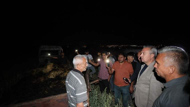 Gece karanlığında, bir grup insan yanmış bitki örtüsü ve enkazın yanında konuşuyor.