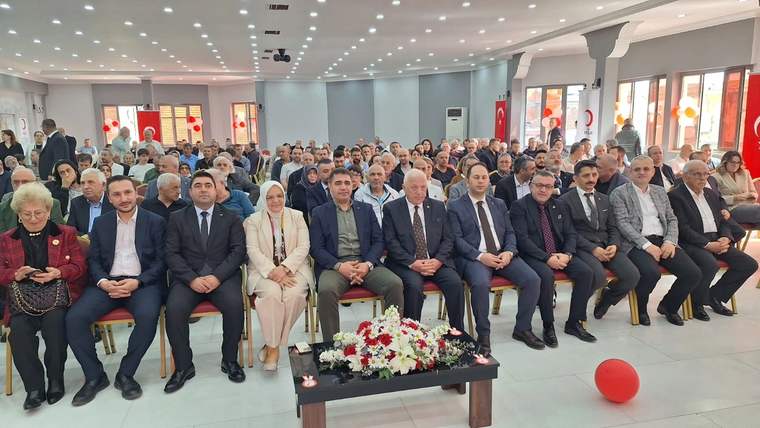 Toplantı salonunda sıralar halinde oturan yetişkinler, önlerinde çiçek aranjmanı ve yerde kırmızı balon görülüyor.