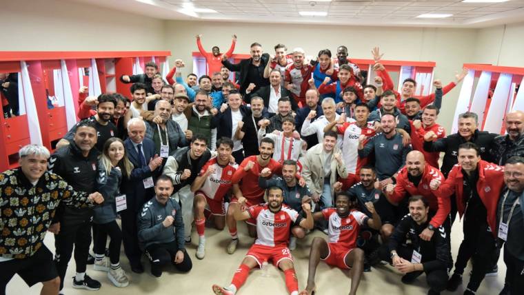 Soyunma odasında kırmızı formalı futbolcular ve teknik ekip, birlikte kameraya sevinçle poz veriyor.