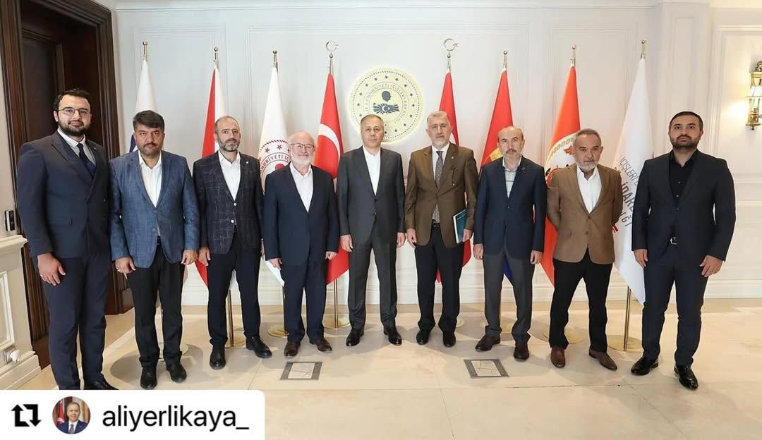 Bir salonda, arkasında Türk bayrakları ve dairesel bir amblem bulunan dokuz erkek yan yana duruyor.
