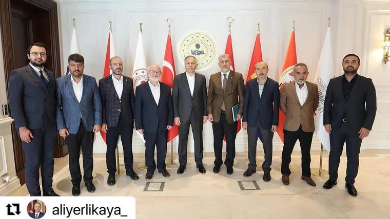 Bir salonda, arkasında Türk bayrakları ve dairesel bir amblem bulunan dokuz erkek yan yana duruyor.