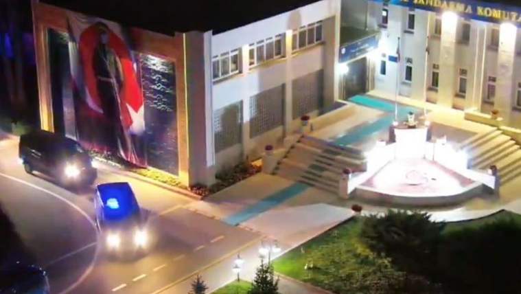 Büyük bir Türk bayrağı ve Atatürk portresi bulunan bir bina önünde, mavi ışıklı bir araç geçiyor.