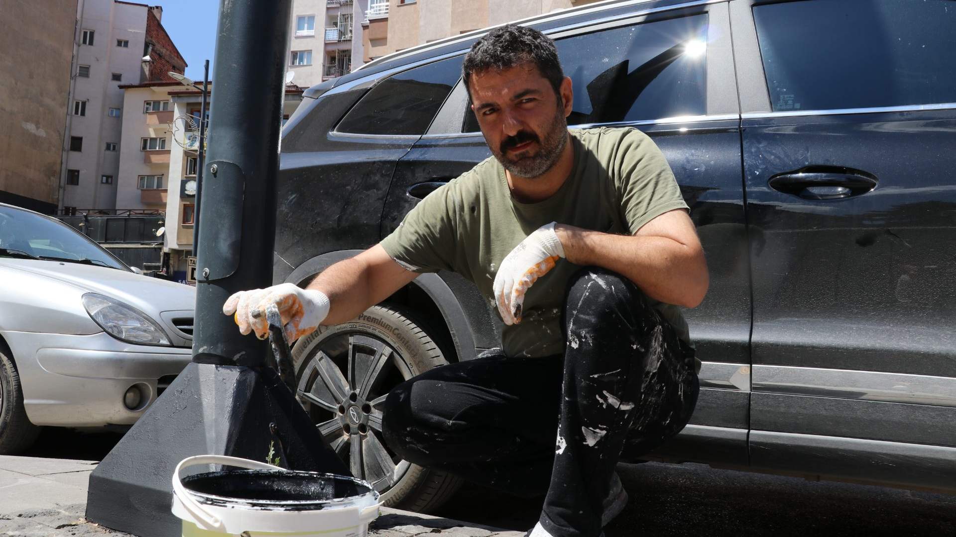 Eldivenli bir kişi, kaldırım kenarındaki siyah direği boya kovasıyla boyarken çömeliyor.