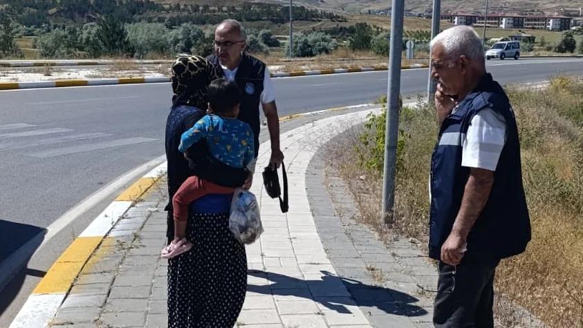 Kaldırımda bir kadın, kucağında çocuk ve elinde poşetle dururken iki yelekli erkek yaklaşıyor.