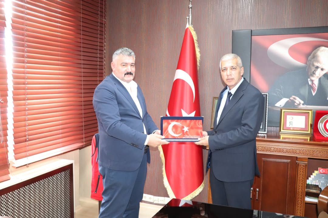 İki kişi, Türk bayrağının önünde üzerinde ay yıldız bulunan bir plaketi birlikte tutuyor.