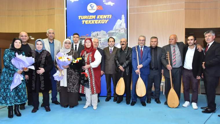 Bir grup insan, sahnede çeşitli müzik aletleriyle poz veriyor, arka planda "Turizm Kenti Tekke köy" yazılı bir afiş var.