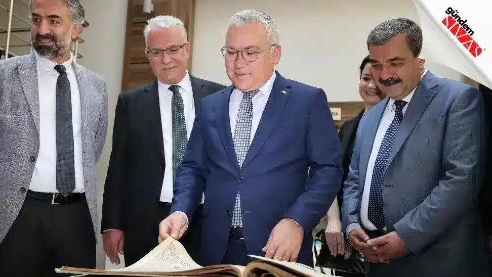 Bir grup insan, büyük bir kitabı inceliyor; ön planda bir kişi kitabı açmış.