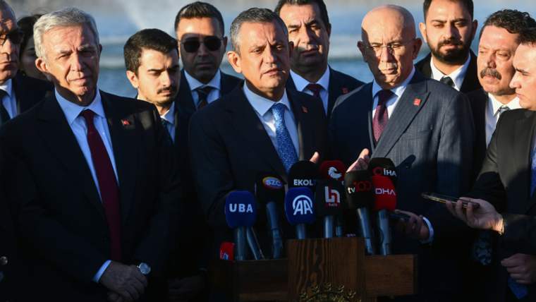 Bir grup erkek, bir podiumun önünde duruyor; podiumda mikrofonlar var ve bir kişi konuşuyor.