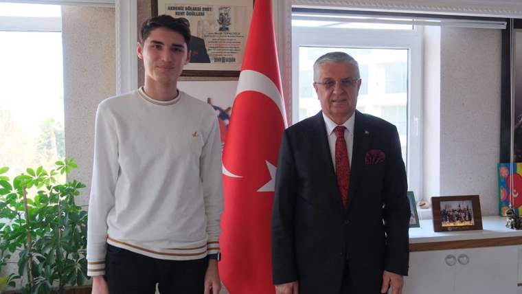 Türk bayrağının önünde, biri genç iki erkek yan yana ayakta duruyor; arkada çerçeveli belgeler bulunuyor.
