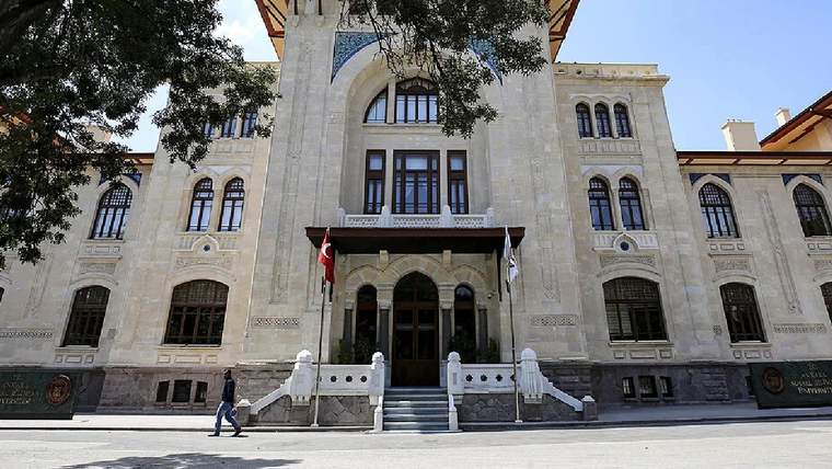 Taş cepheli, kemerli pencereleri ve girişinde Türk bayrakları bulunan üç katlı bir bina; önünden bir kişi geçiyor.