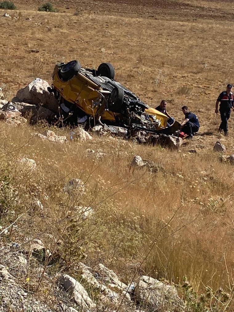 Kuru otlarla kaplı arazide ters dönmüş sarı bir araç ve çevresinde duran üç kişi görülüyor.