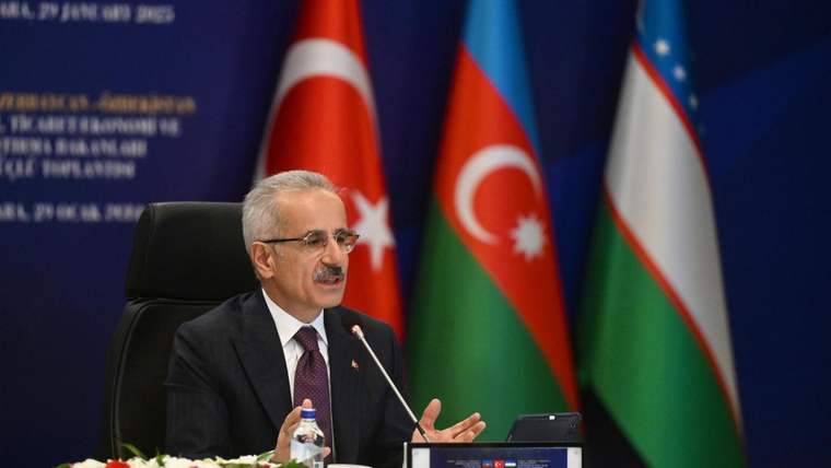 Mikrofon başında konuşan bir kişi, masada oturuyor; arkasında Türkiye, Azerbaycan ve Özbekistan bayrakları görülüyor.
