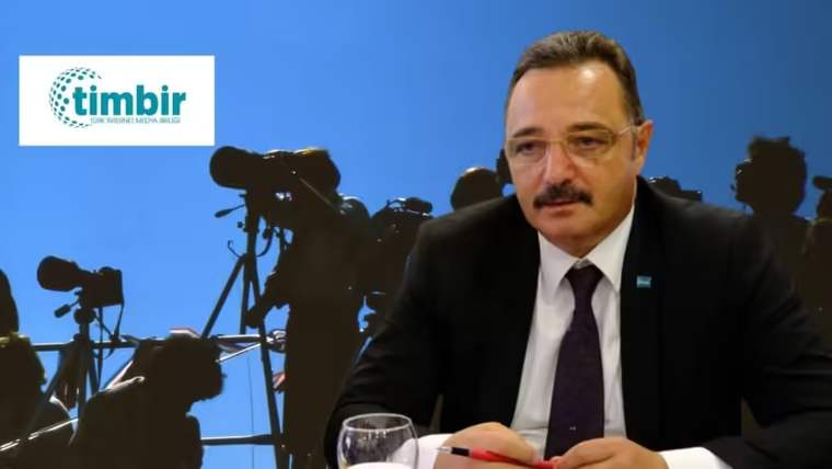 Bir kişi, masada oturmuş, kameraların siluetlerinin olduğu arka planda konuşuyor.