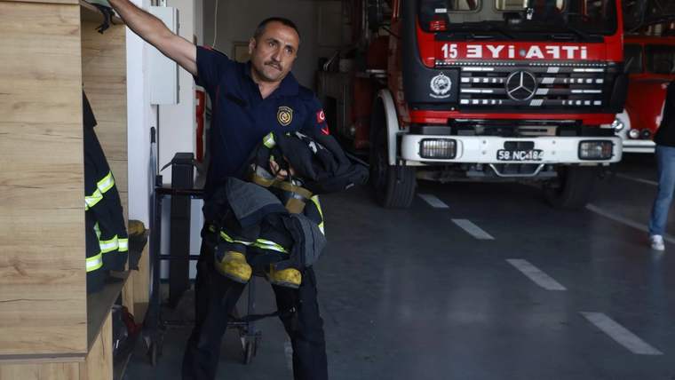 İtfaiye garajında bir itfaiyeci, yanında duran kırmızı itfaiye aracının önünde koruyucu kıyafet tutuyor.