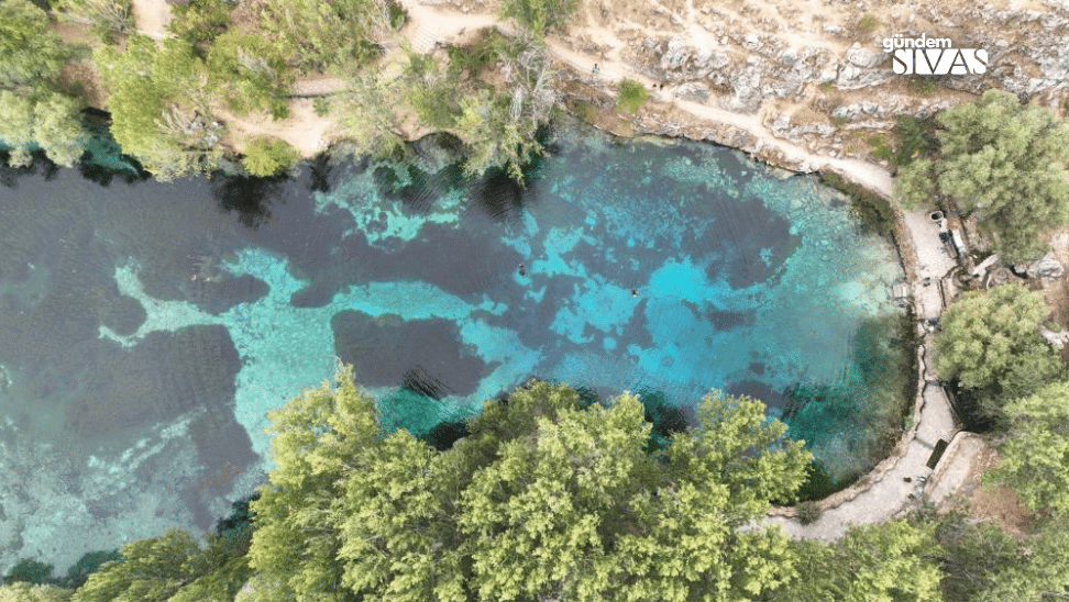 Suyun çeşitli tonlarında mavi ve yeşil renkler taşıdığı bir gölet, çevresinde ağaçlar ve kayalıklar var.