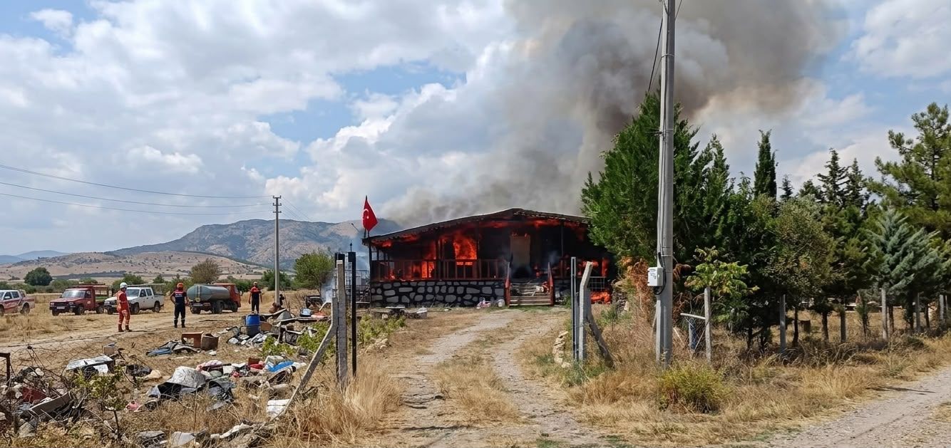 Ağaçların yanındaki tek katlı yapı alevler içinde; çatısından yoğun siyah duman yükseliyor.