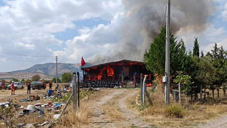 Ağaçların yanındaki tek katlı yapı alevler içinde; çatısından yoğun siyah duman yükseliyor.