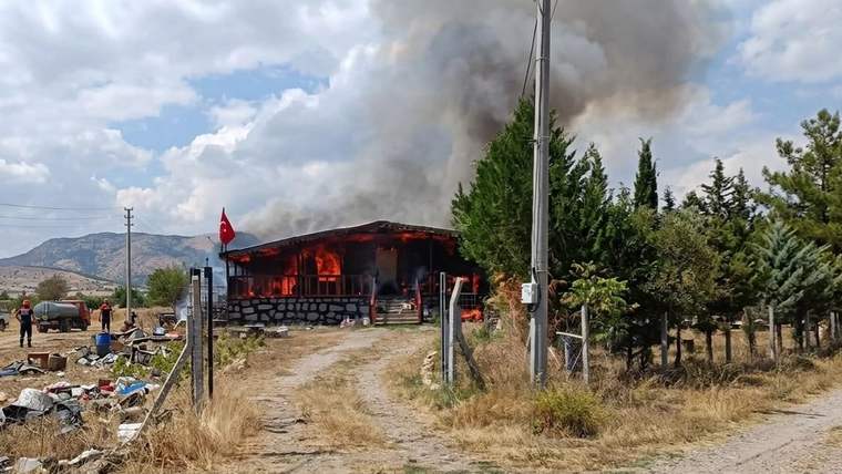 Ağaçların yanındaki tek katlı yapı alevler içinde; çatısından yoğun siyah duman yükseliyor.