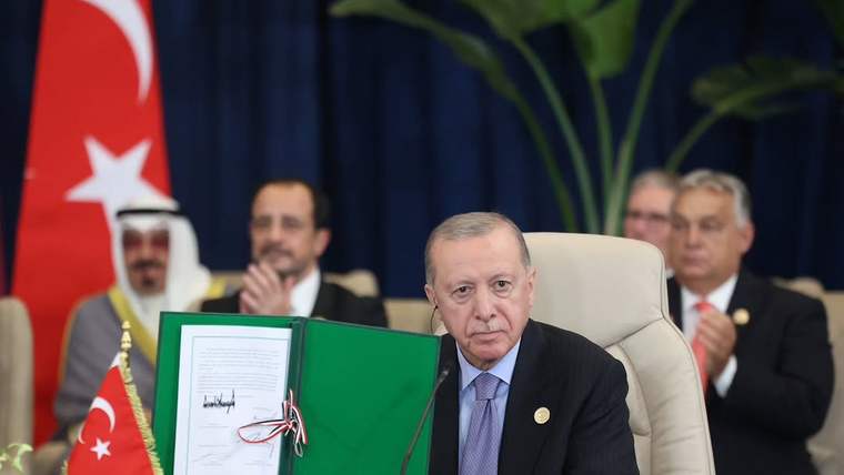 Masa başında oturan bir kişi, kırmızı kurdeleli ve mühürlü bir belgeyi açık halde tutuyor; arkada bayraklar var.