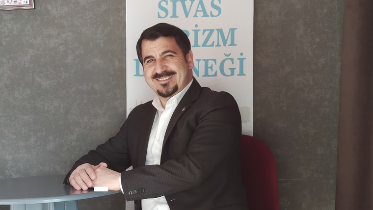 Masada oturan gülümseyen bir kişi, arkasındaki afişte kısmen görülen “Sivas Turizm Derneği” yazısının önünde duruyor.