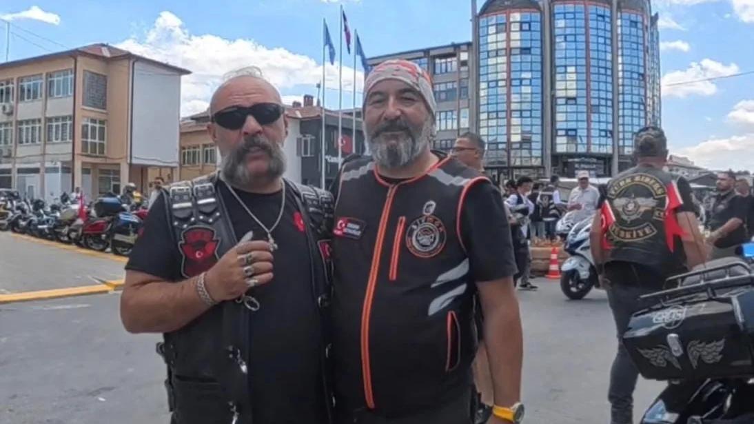 Motosiklet giysileri giymiş iki adam, gülümseyerek yan yana duruyor.