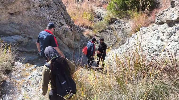Kayalık ve kuru otlarla çevrili arazide sırt çantalı dört kişi ilerlerken görülüyor.