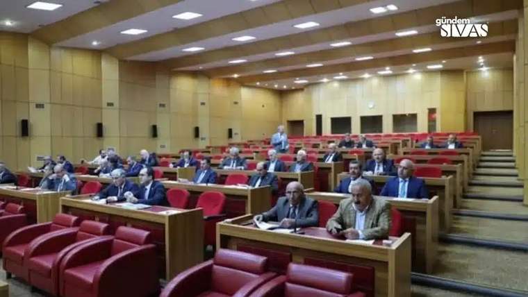 Ahşap sıralarla düzenlenmiş salonda çok sayıda kişi oturuyor; ön sırada iki kişi görülüyor.