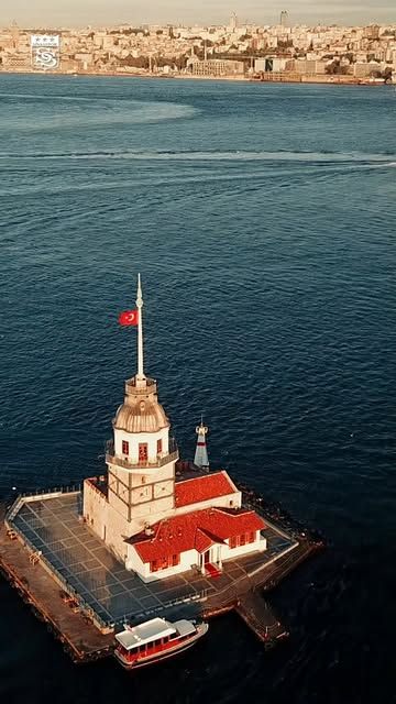 Denizin ortasındaki küçük adacıkta Türk bayraklı, kırmızı çatılı tarihi bir kule ve yanında tekne görülüyor.